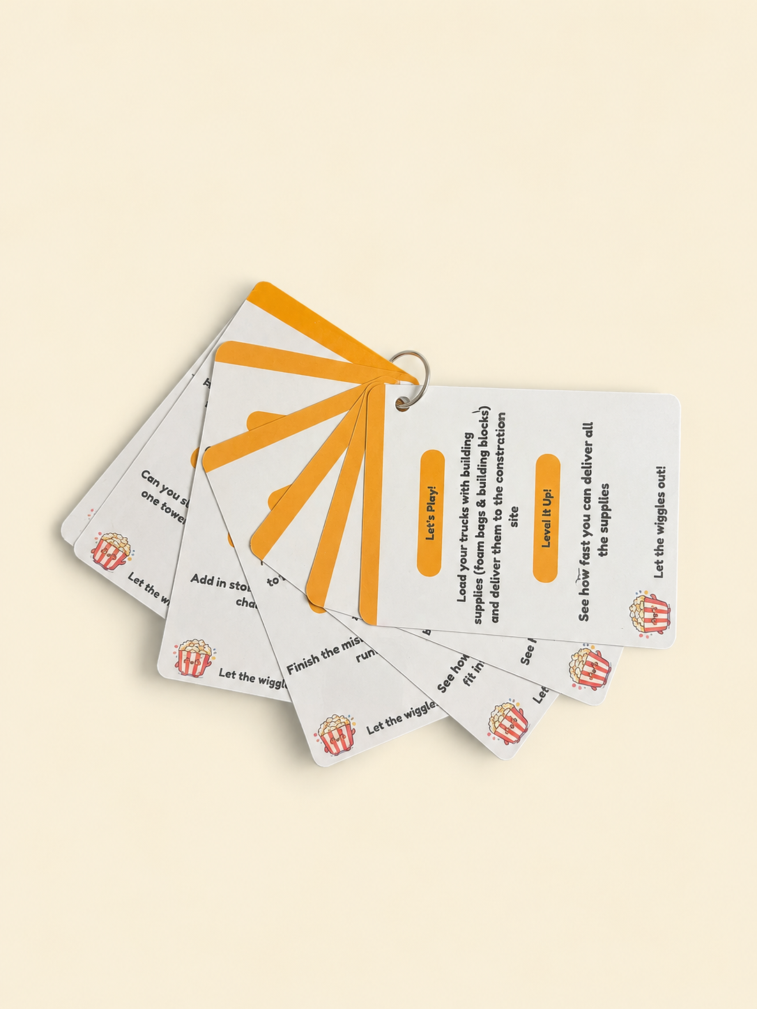 A fanned-out set of small instructional cards on a plain warm beige background. The cards are white with orange and black text, plus small illustrated icons, construction themed play prompts