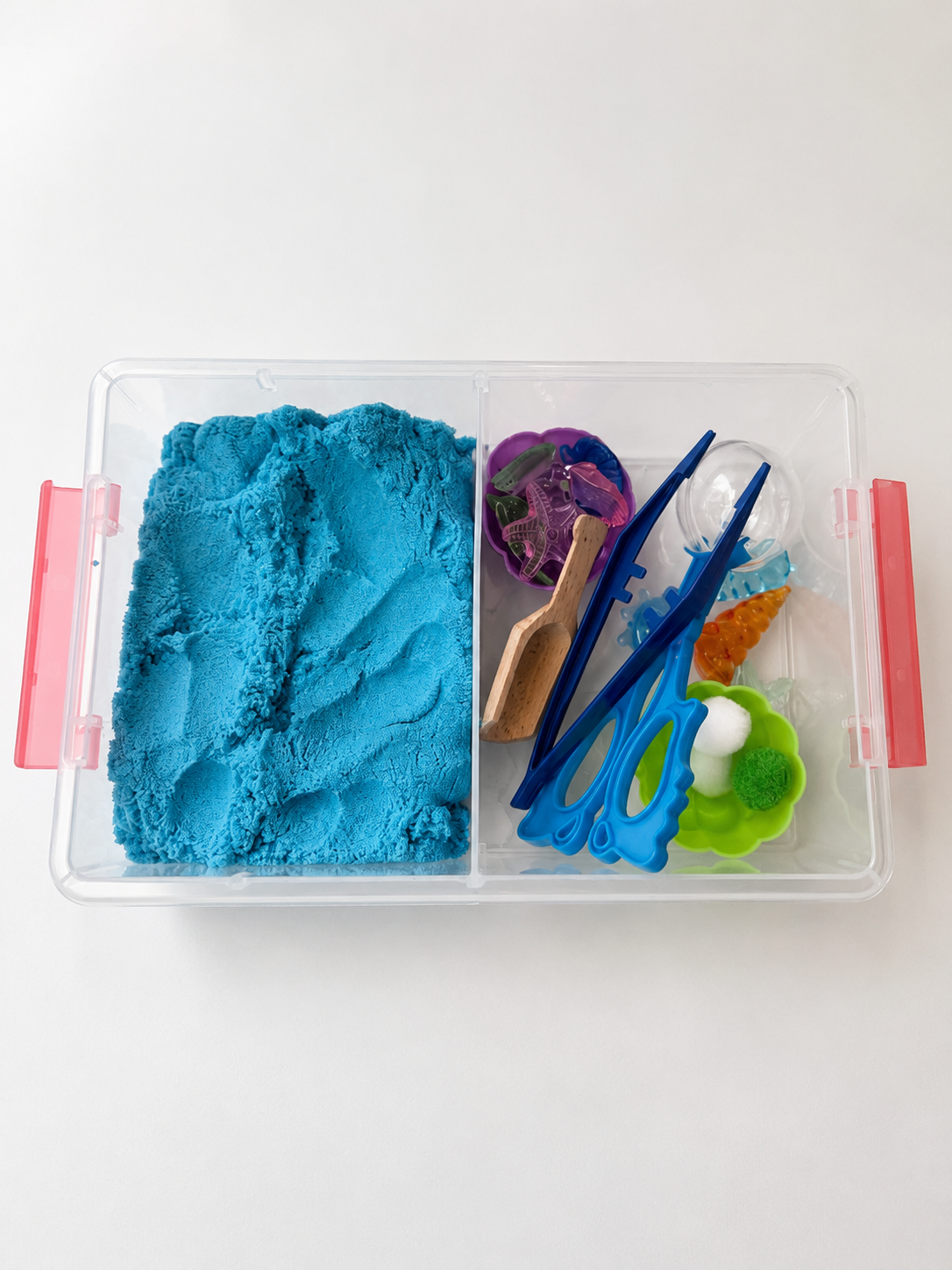 A clear plastic divided sensory play box with pink side handles, filled with bright blue kinetic sand in one compartment and small colorful toy tools in the other, photographed on a plain white background.