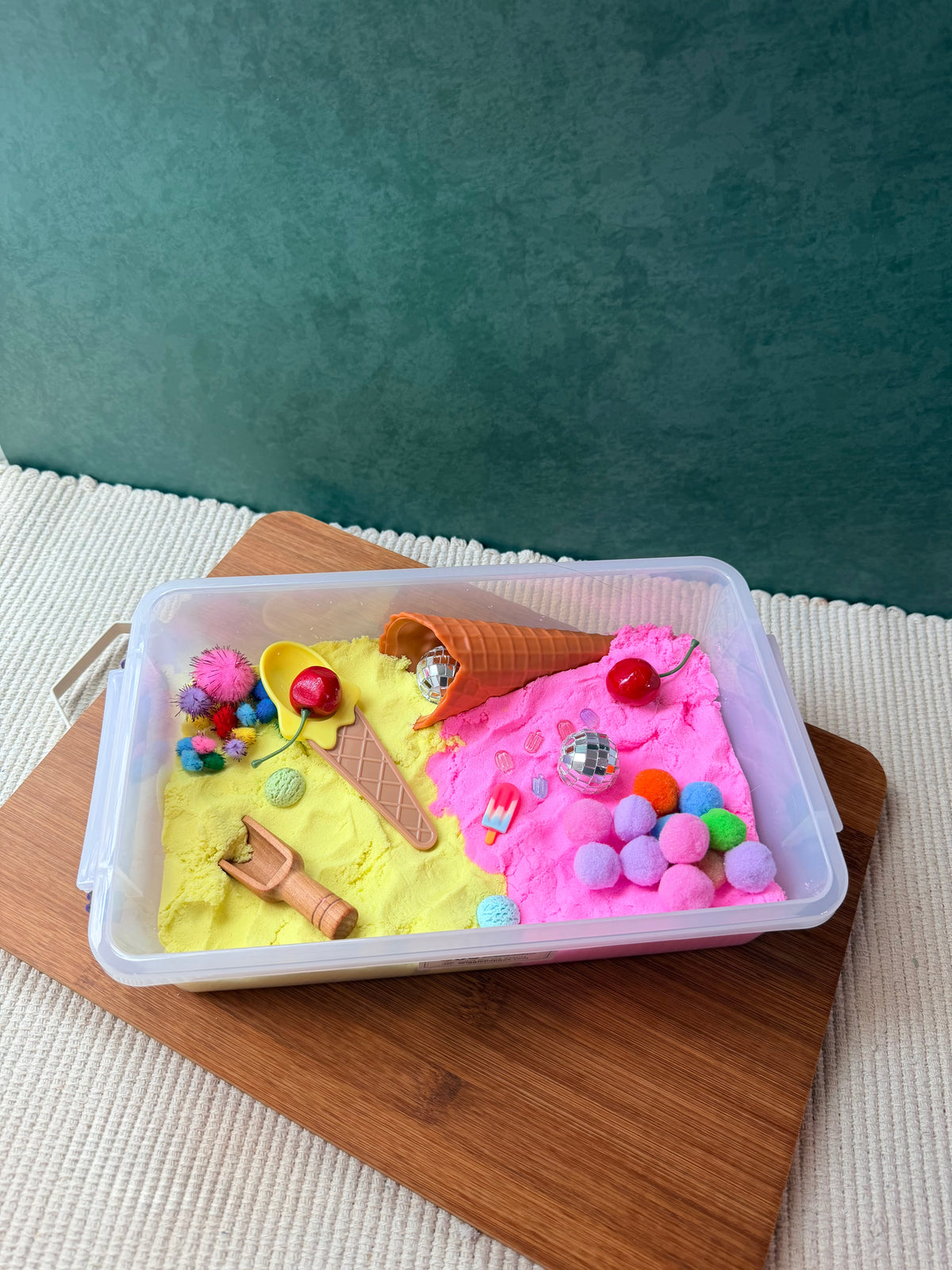 A clear plastic sensory tray filled with split sand in bright yellow and pink, decorated with miniature ice cream cones, colorful pom-poms, beads, and small toy accessories, displayed on a wooden board against a teal wall.