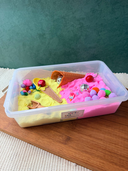 A clear plastic sensory bin filled with split sand in pastel yellow and pink, decorated with miniature ice cream cones, colorful pom-poms, beads, and small toy accessories, set on a wooden board against a dark green textured background.