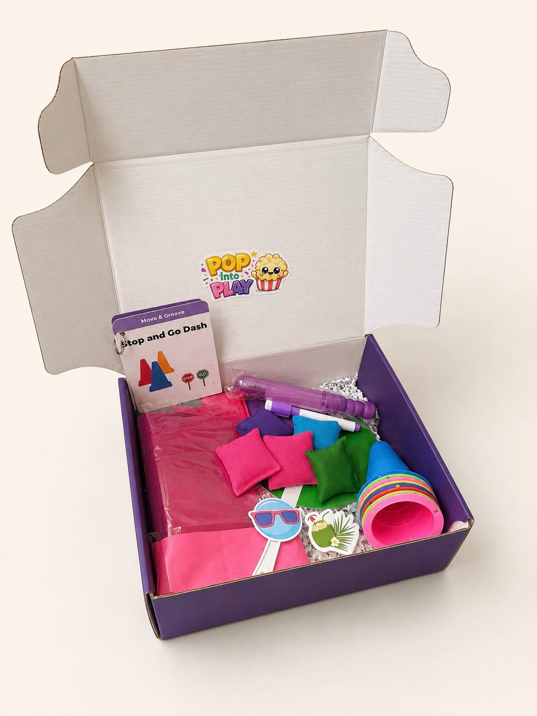 An open purple cardboard gift box containing a Pop Up Party Play kit with various colorful play accessories including toss bean bags, bubble wand, cones, stickers, dry erase marker, stop and go sign, including pink and green fabric pieces,  and a small instruction card.