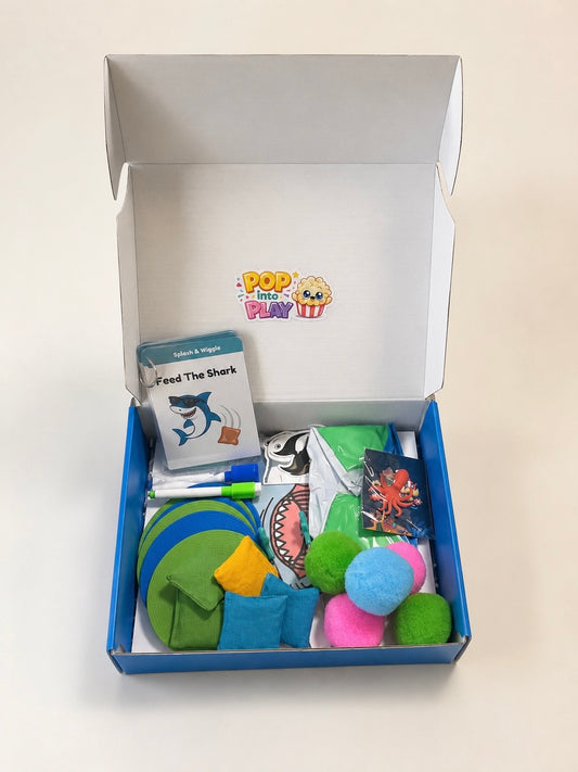 An open blue cardboard gift box on a white background, filled with a children's play set including a shark-themed card, plush toys, and colorful accessories. The inside lid shows the 'Pop It Play It' logo.