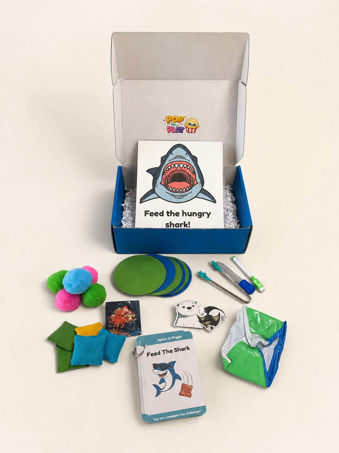 A children's educational toy set titled 'Feed the Hungry shark!' arranged on a light beige background. The set includes a blue and white storage box with a shark-themed card, green circular pieces, colorful pom-poms, small fabric pieces, a pen-like tool, and other accessories.