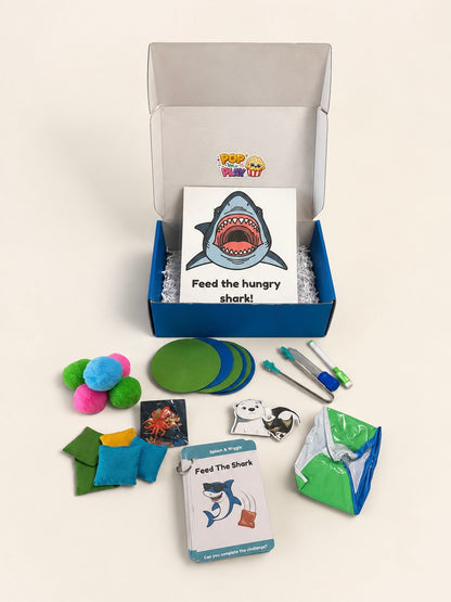 A children's educational toy set titled 'Feed the Hungry shark!' arranged on a light beige background. The set includes a blue and white storage box with a shark-themed card, green circular pieces, colorful pom-poms, small fabric pieces, a pen-like tool, and other accessories.