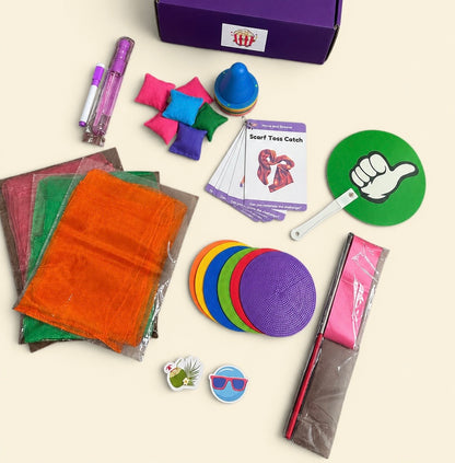 A flat-lay view of a dance activity kit containing multiple items including a purple box, colored fabric squares, cones, a play prompt activity cards, a green thumbs-up sign, and small accessories on a light beige background.