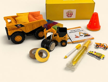 A children's construction-themed toy set arranged on a light beige background, including a yellow dump truck with play bean bag toss, a small yellow construction vehicle, a roll of road tape, a cone, a marker, a bubble wand, small vehicle stickers, a booklet of activity card play prompts, and a yellow gift box.