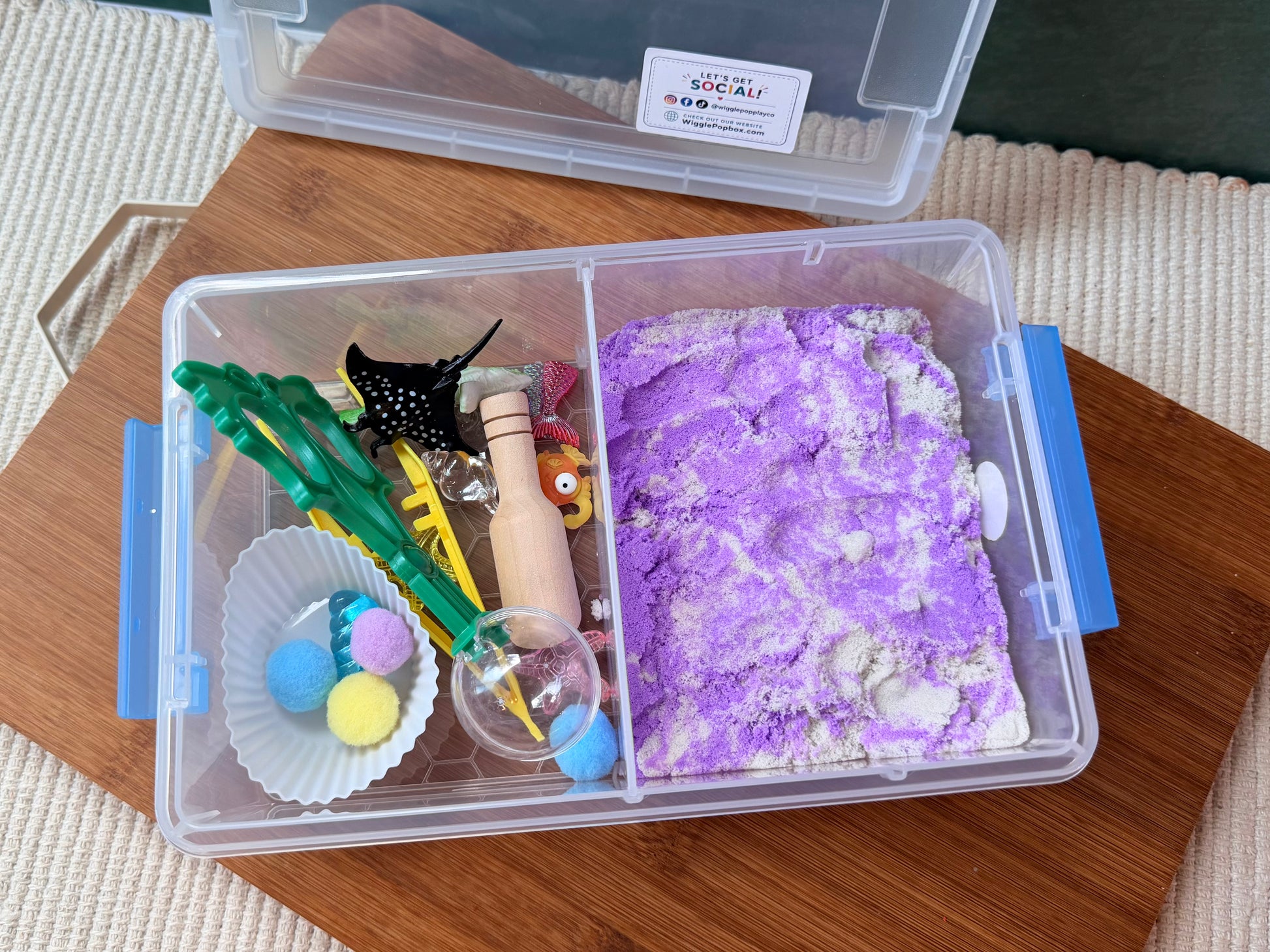 A clear plastic sensory bin organizer with blue latches, shown open on a wooden tray. The left compartment contains colorful sensory toys including green tongs, a wooden bead, and pom-poms, while the right compartment holds a purple and white textured sand-like material.