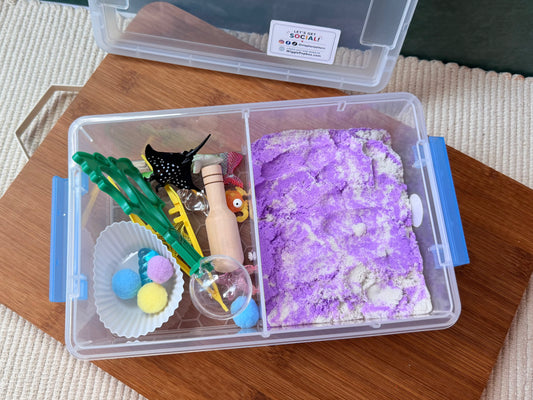 A clear plastic sensory bin organizer with blue latches, shown open on a wooden tray. The left compartment contains colorful sensory toys including green tongs, a wooden bead, and pom-poms, while the right compartment holds a purple and white textured sand-like material.