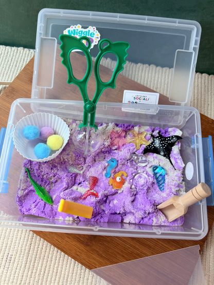 An open clear plastic sensory bin filled with purple kinetic sand and surrounded by small toys and accessories, including green scissors, pom-poms, shells, and a wooden stirrer, photographed on a tabletop.