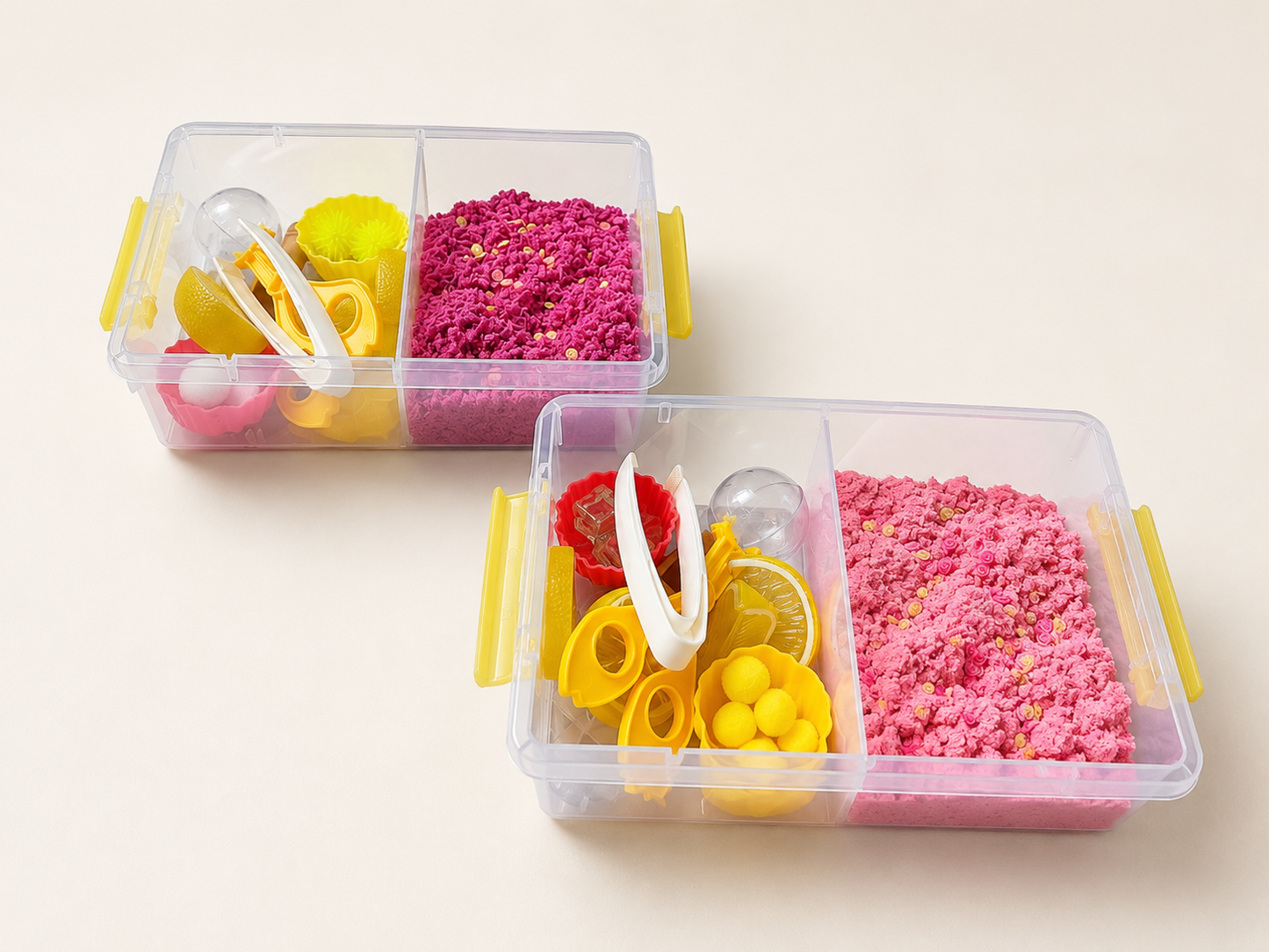 Two clear plastic sensory bins with yellow handles, filled with bright pink sand and colorful toy accessories, on a light beige background.