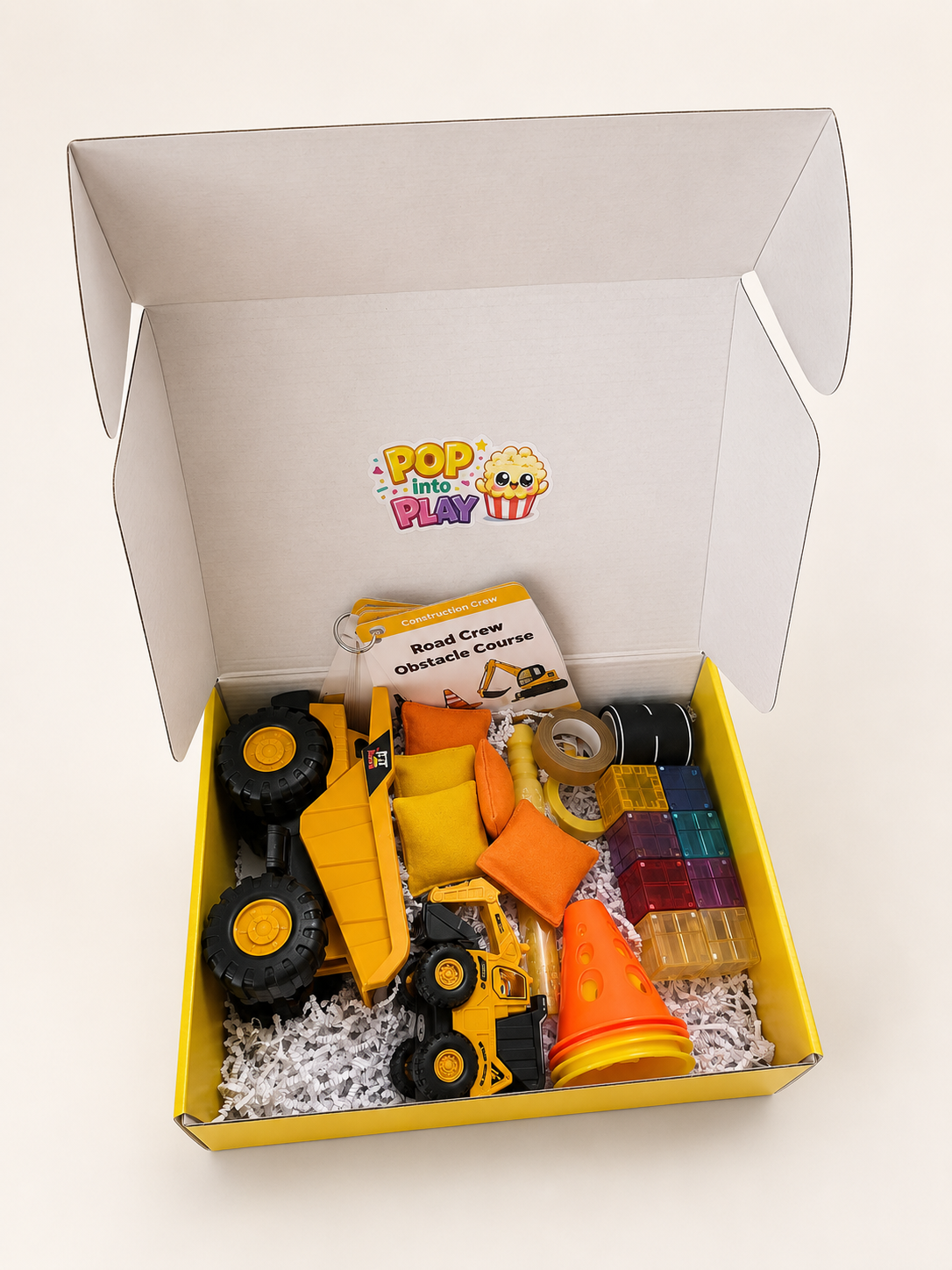A children's construction-themed gift box from wigglepop play co, containing a yellow toy dump truck, a small yellow construction vehicle, construction cones, translucent magnetic building block pieces, and a activity cards with play prompts, all arranged in white shredded paper inside a yellow and white box. Done for you play delivered to your door. 