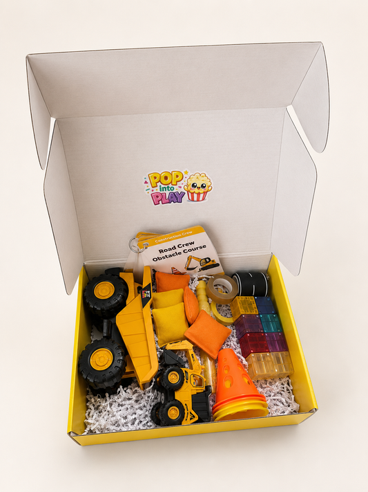 A children's construction-themed gift box from wigglepop play co, containing a yellow toy dump truck, a small yellow construction vehicle, construction cones, translucent magnetic building block pieces, and a activity cards with play prompts, all arranged in white shredded paper inside a yellow and white box. Done for you play delivered to your door. 