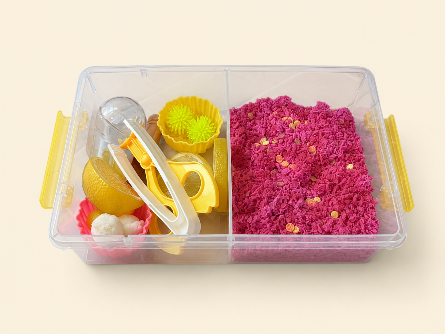A clear plastic divided storage container with yellow handles, shown on a light beige background. The right compartment contains bright pink play foam with small lemons, and the left compartment holds yellow and white sensory play tools and lemonade themed play pieces.