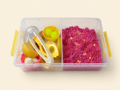A clear plastic divided storage container with yellow handles, shown on a light beige background. The right compartment contains bright pink play foam with small lemons, and the left compartment holds yellow and white sensory play tools and lemonade themed play pieces.