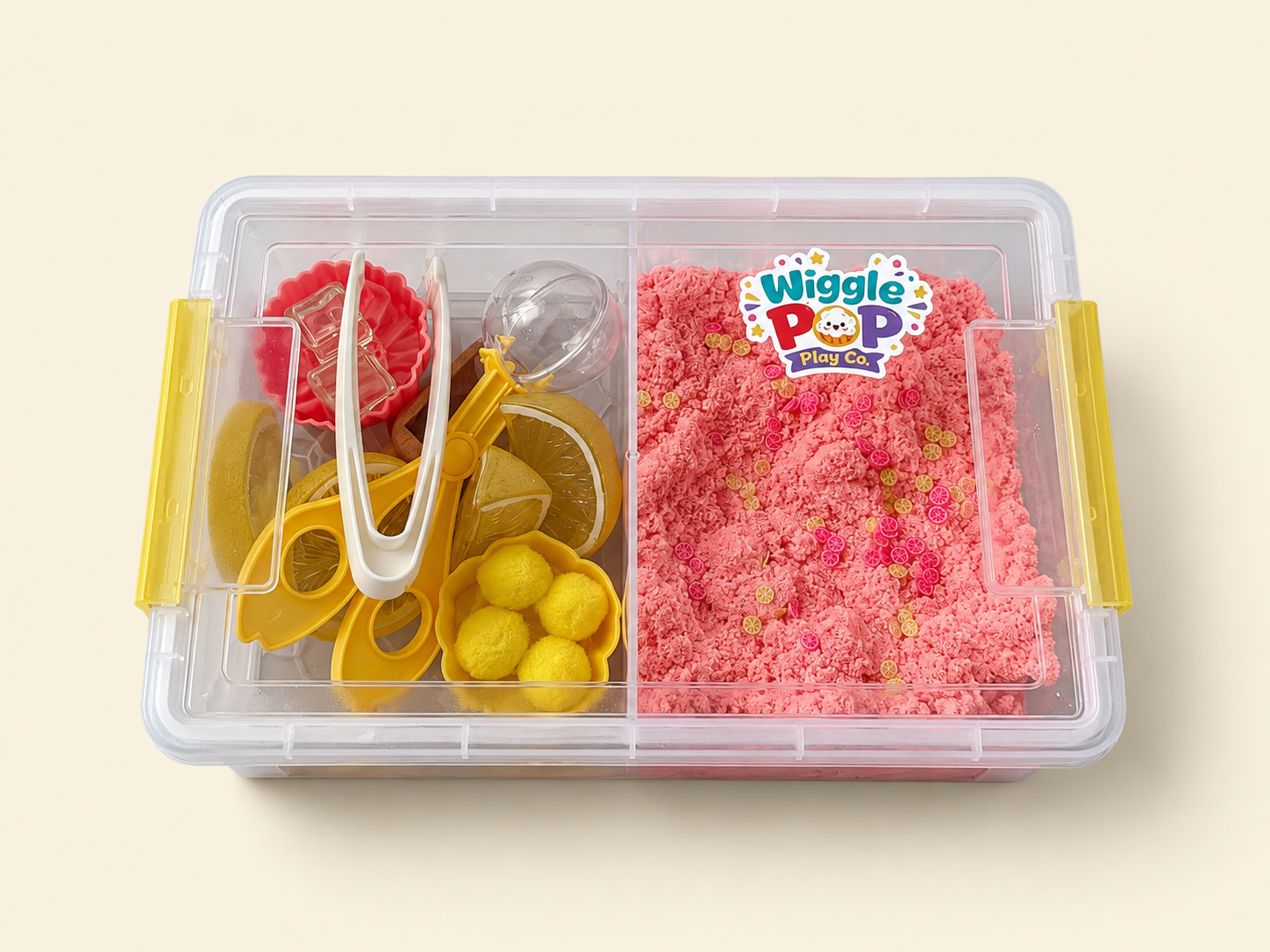 A clear plastic storage container with yellow handles, divided into two sections. The left side contains yellow and white toy sensory play accessories, and the right side is filled with pink kinetic sand.