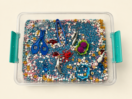 An overhead view of a clear plastic sensory bin with teal handles, filled with blue glitter, colorful beads, and small toy-like accessories including blue scissors, a red crab, a purple flower, and silver fish, set against a light beige background.