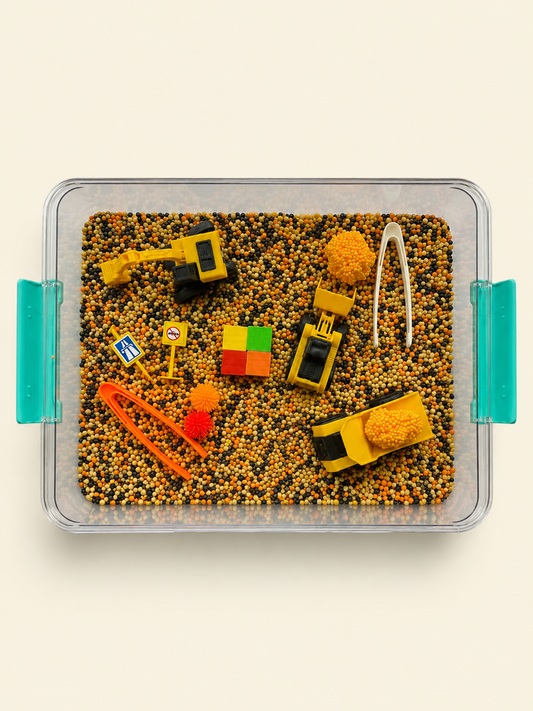 A clear plastic sensory tray filled with small multicolored beads and decorated with construction-themed toys, including yellow toy vehicles, an orange tool, a white trowel, and a small multicolor cube, shown on a light beige background.