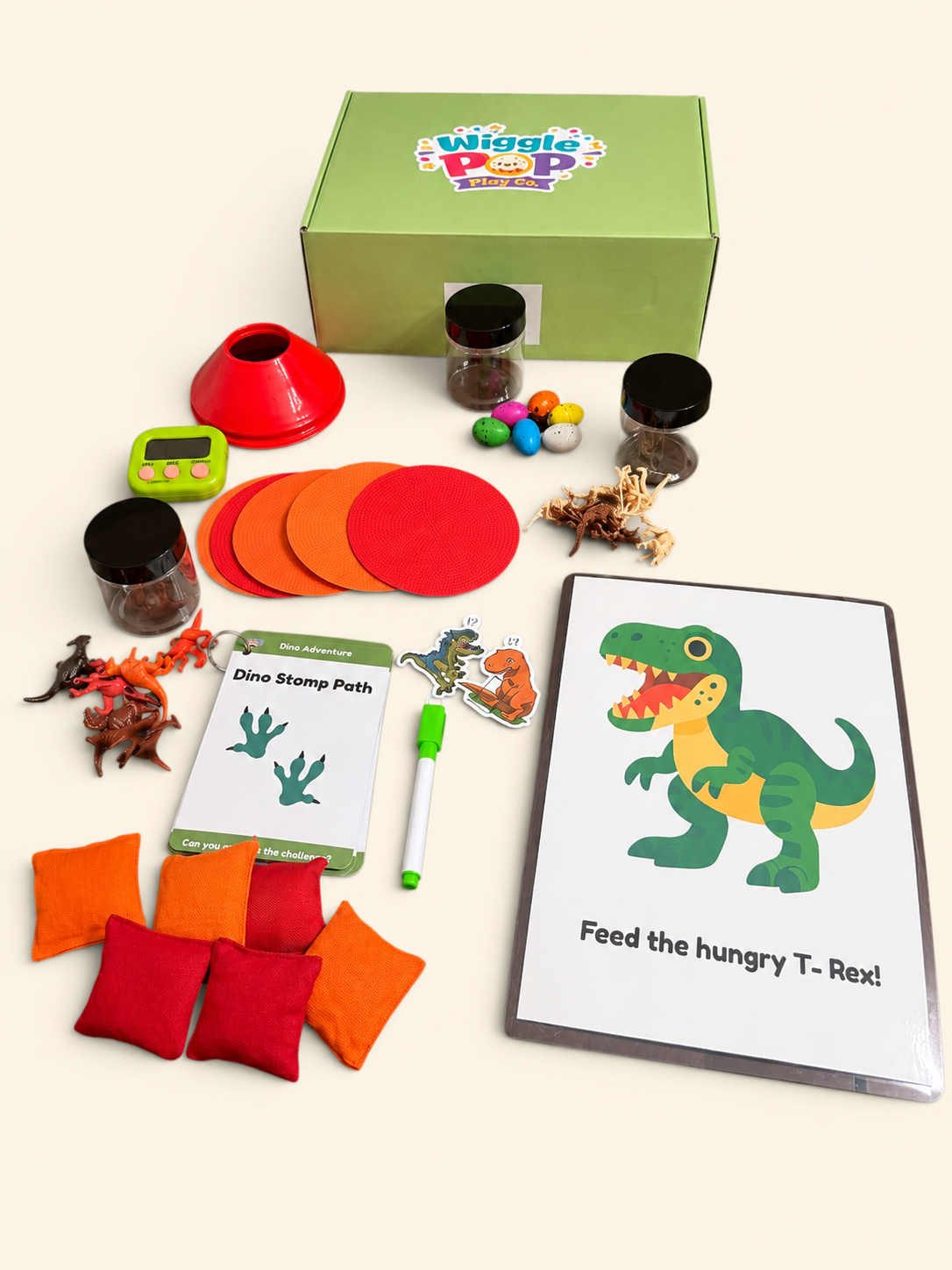 A children's educational dinosaur activity kit laid out on a light cream background, including a green box with the Wiggle Pop Dinosaur logo, a green T-rex activity board reading 'Feed the hungry T-Rex!', red and orange fabric discs, small jars, toy dinosaurs, a stamp path card, a marker, and bean bags.