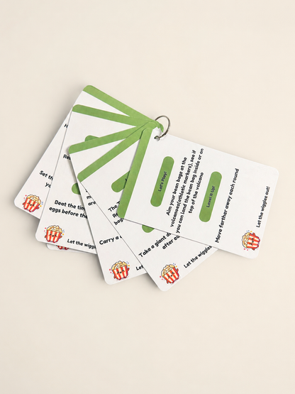 A set of printed cards tied together with a green ribbon on a metal ring, photographed on a plain light beige background. The cards are white with green and black text and small red-and-yellow popcorn graphics.