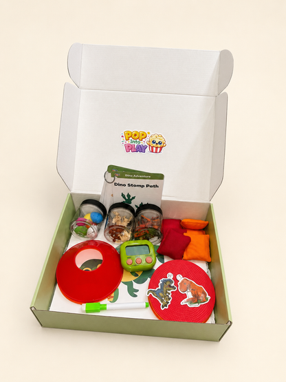 An open white cardboard gift box containing a dinosaur-themed sensory play kit. The kit includes a green and white 'Dino Stomp Path' card, a green handheld timer, a red circular plastic piece, a red stamp pad with a dinosaur illustration, two small clear cups filled with colorful pieces, and two small orange and red fabric pieces.