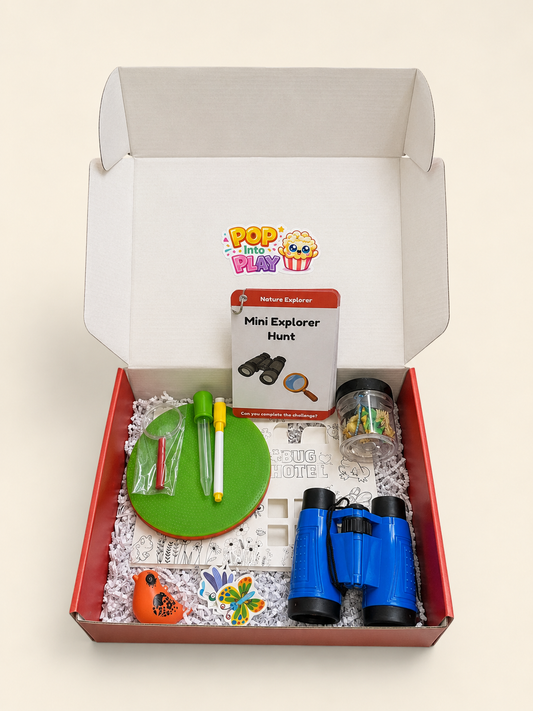 An open white cardboard gift box with a red inner tray, filled with a children's nature explorer kit on white paper shred. The kit includes blue binoculars, floor dots, a yellow dry erase marker, a green squeeze syringe, a small orange bird whistle, a clear jar with play bugs, and a card that reads 'Mini Explorer Hunt.