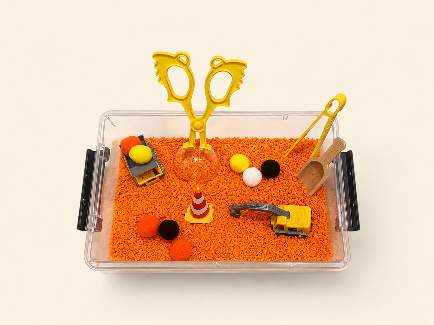 A clear plastic sensory bin filled with bright orange play foam and small construction-themed toys, including yellow toy scissors, a small excavator, a traffic cone, pom-poms, and a wooden scoop, photographed on a light cream background.