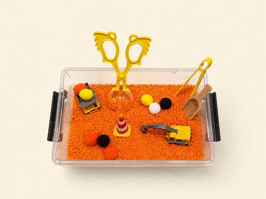 A clear plastic sensory bin filled with bright orange play foam and small construction-themed toys, including yellow toy scissors, a small excavator, a traffic cone, pom-poms, and a wooden scoop, photographed on a light cream background.