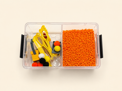 A clear plastic divided storage box with black side handles, shown from above on a light beige background. The left compartment contains small yellow construction tools and assorted pom-poms, while the right compartment is filled with bright orange play foam material.
