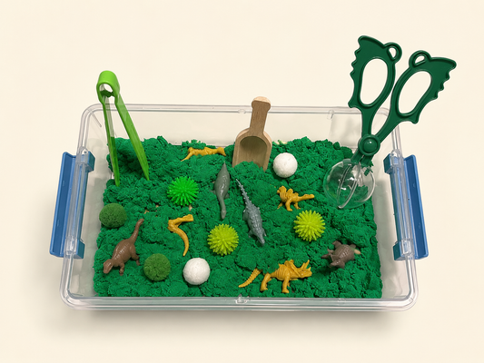 A clear plastic sensory tray filled with green kinetic sand and small toy dinosaurs, plants, a scoop, and green-handled tongs and scissors, photographed on a plain light beige background.