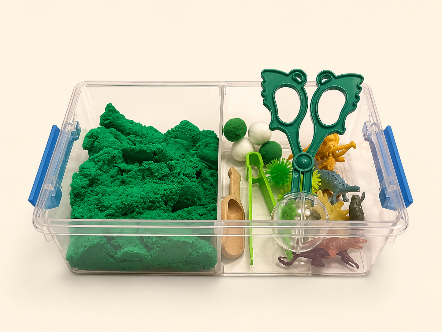 A clear plastic divided storage bin with blue handles, filled with green kinetic sand in one compartment and small toy-like play accessories in the other, photographed on a plain light background.