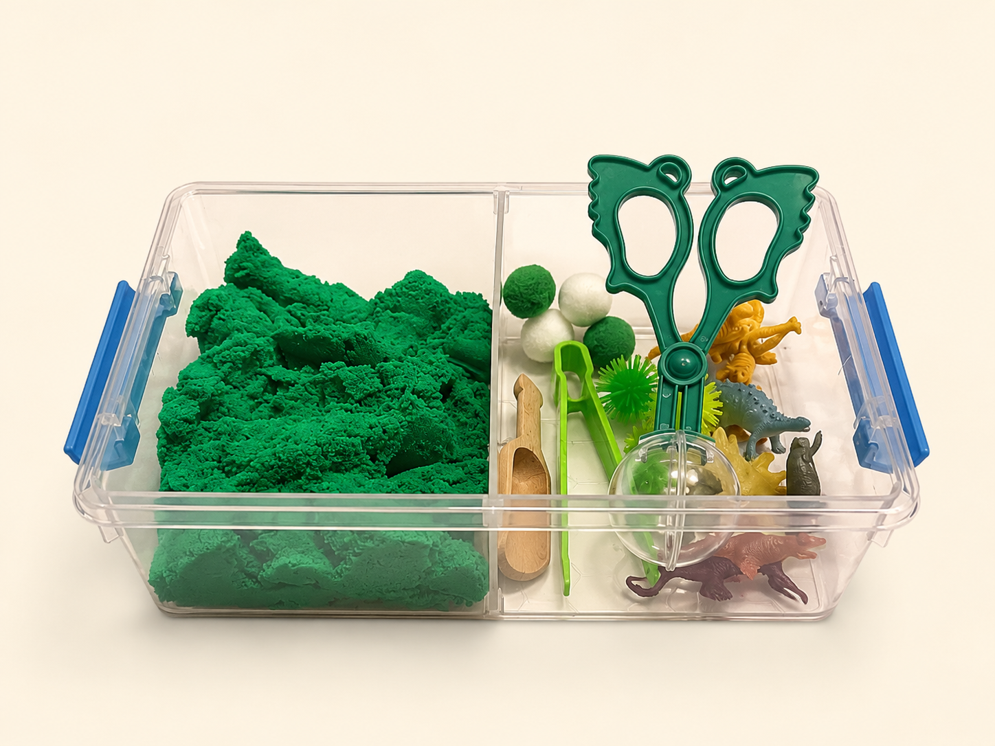 A clear plastic divided storage bin with blue handles, filled with green kinetic sand in one compartment and small toy-like play accessories in the other, photographed on a plain light background.