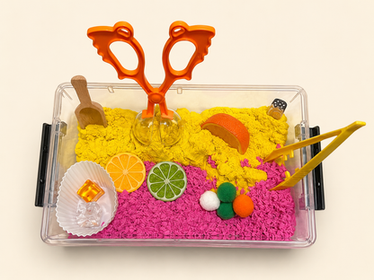 A clear plastic sensory tray filled with bright pink play foam and yellow kinetic sand, decorated with colorful toy fruits, a wooden scoop, orange plastic scissors, and yellow tongs, photographed on a plain light background.
