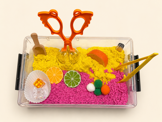 A clear plastic sensory tray filled with bright pink play foam and yellow kinetic sand, decorated with colorful toy fruits, a wooden scoop, orange plastic scissors, and yellow tongs, photographed on a plain light background.