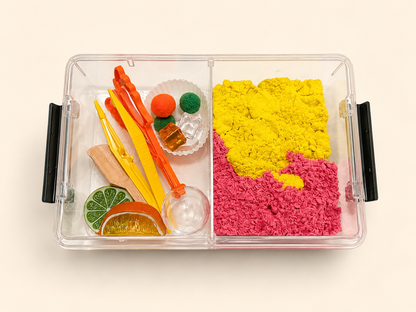 A clear plastic divided sensory box with black side handles, filled with bright play materials: a large yellow and pink sand-like section on the right, and orange, yellow, green, and brown toy accessories on the left, photographed on a plain light background.