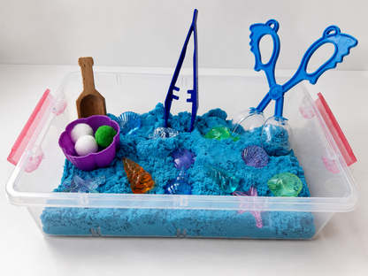 A clear plastic sensory bin filled with bright blue kinetic sand and decorated with toy sea-themed items including a purple shell bowl with white and green balls, a wooden scoop, blue fishing rods, blue tongs, and translucent plastic shells.