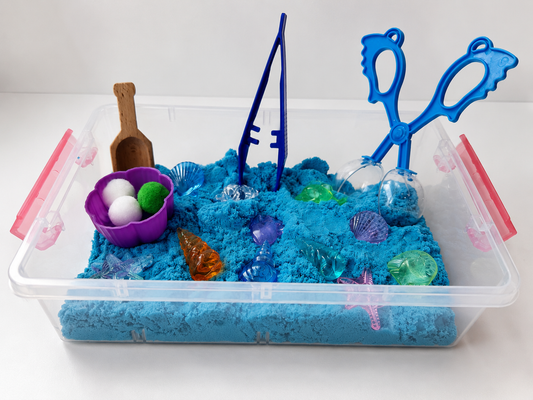 A clear plastic sensory bin filled with bright blue kinetic sand and decorated with toy sea-themed items including a purple shell bowl with white and green balls, a wooden scoop, blue fishing rods, blue tongs, and translucent plastic shells.