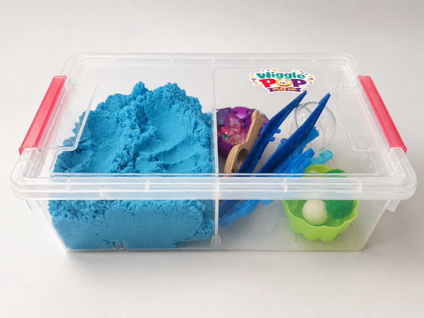 A clear plastic divided storage bin with pink handles, containing bright blue kinetic sand and several small toy tools, photographed on a white background.