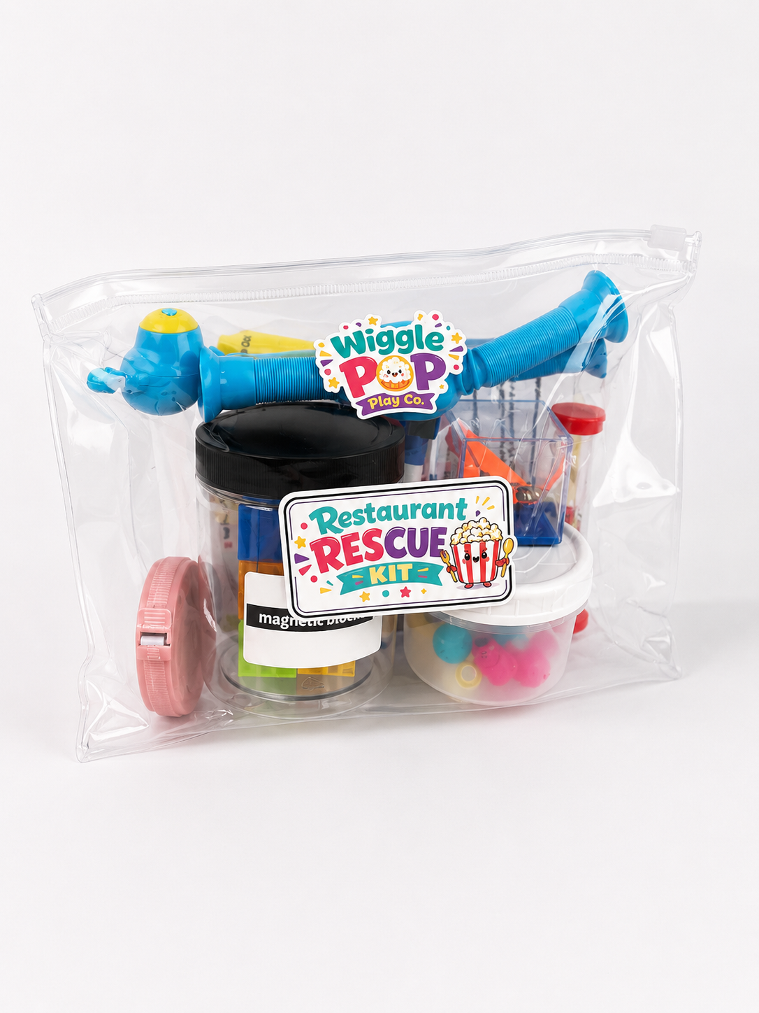 A toy restaurant rescue kit in a clear zippered bag, featuring a blue telescopic toy, a tape measure, a small challenge cube, and containers with colorful magnetic pieces. The front has a 'Wiggle Pop' logo and a 'Restaurant Rescue' sticker.